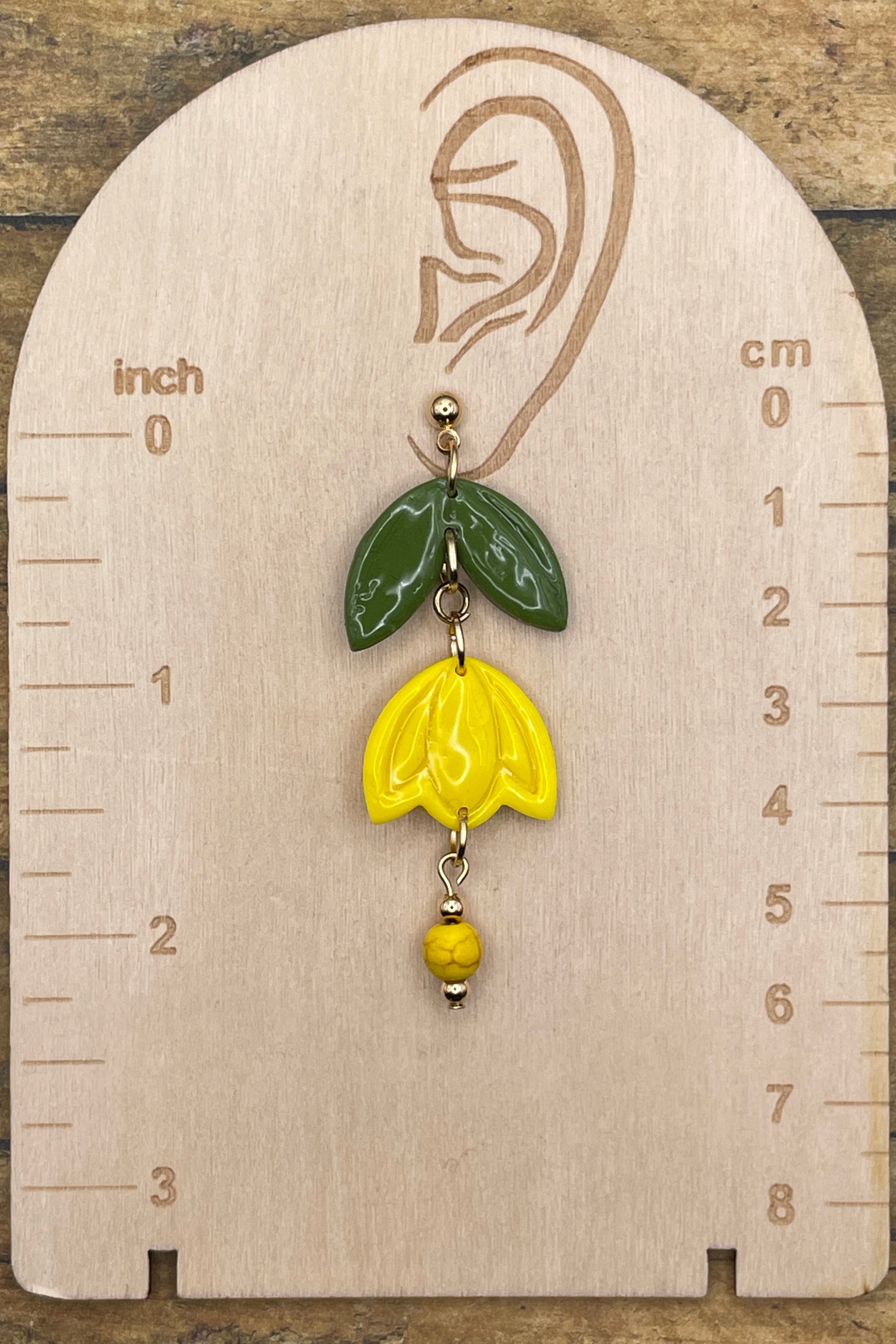Yellow flower-shaped earring on a wooden measurement card with inch and cm scales.