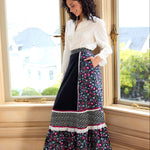 Woman standing in a room with large windows, wearing a white blouse and a long floral skirt.