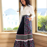 Woman standing in a room with large windows, wearing a white blouse and a long floral skirt.