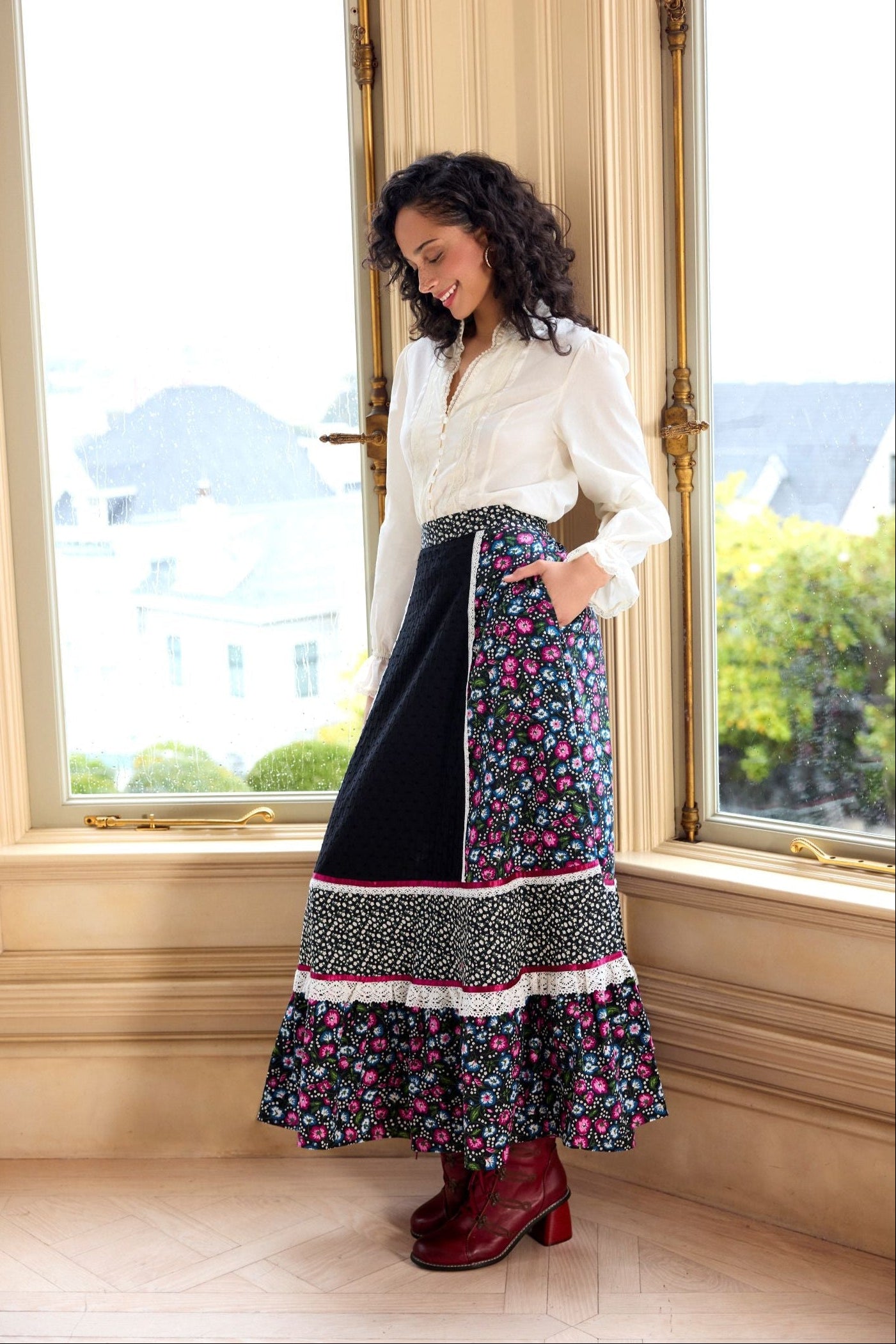 Woman standing in a room with large windows, wearing a white blouse and a long floral skirt.
