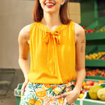 Woman wearing a yellow top and floral skirt with a fruit-themed pattern, standing in front of a produce stand.