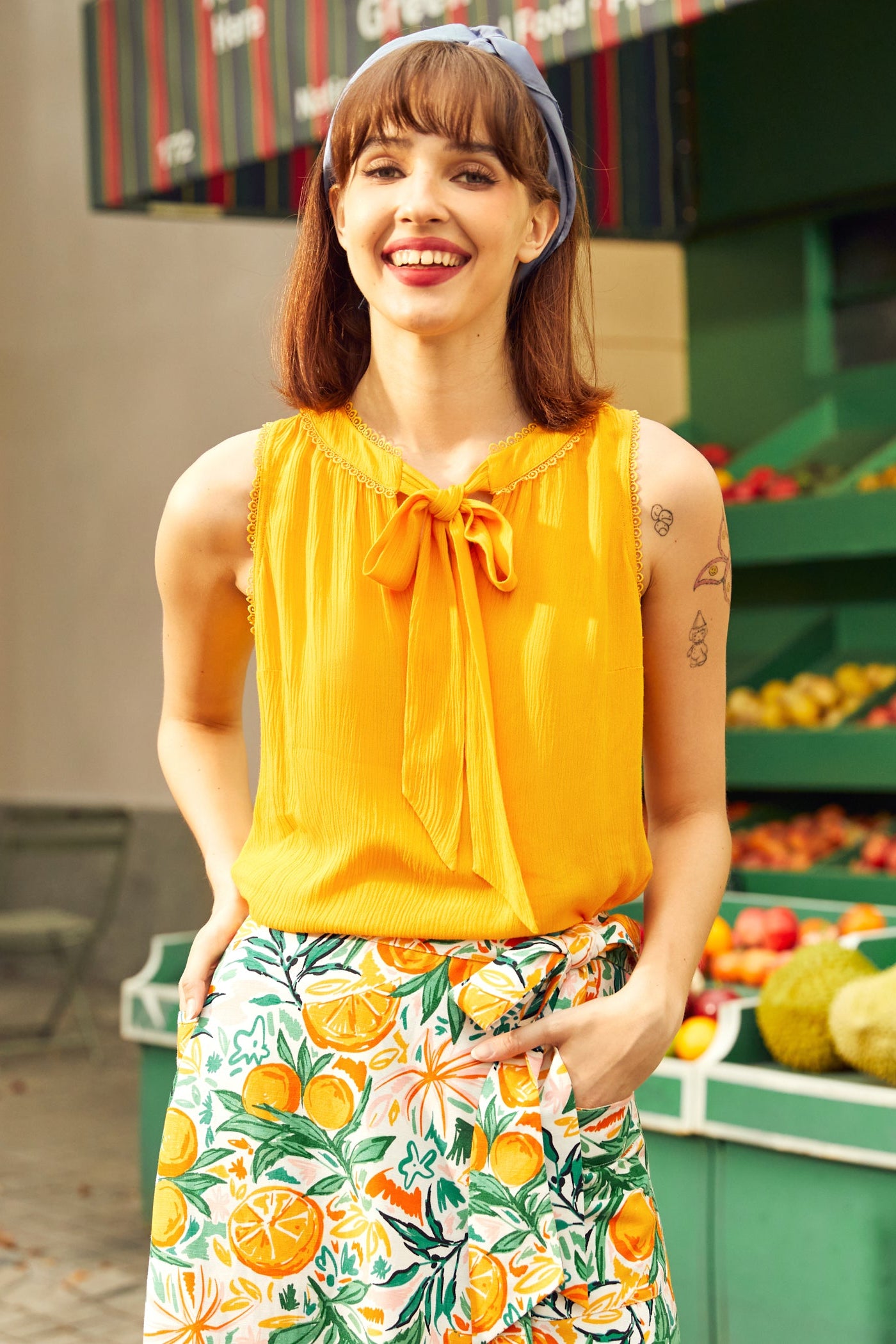 Woman wearing a yellow top and floral skirt with a fruit-themed pattern, standing in front of a produce stand.