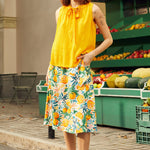 Woman in a yellow top and floral skirt standing in front of a fruit stand.
