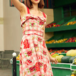 Woman in a pink floral summer dress standing in front of a fruit stand.