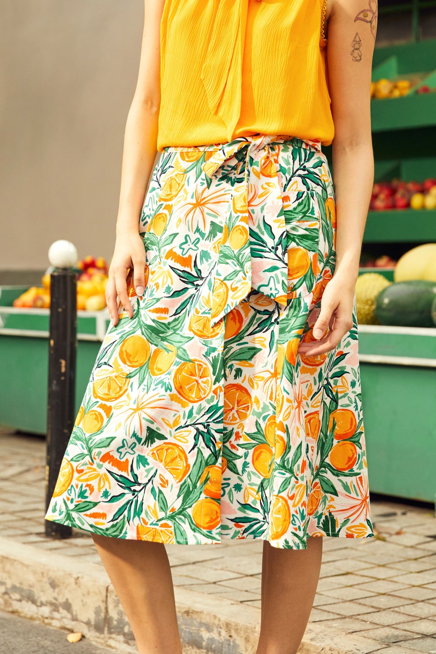 Person wearing a yellow top and a colorful wrap skirt with citrus fruit pattern outdoors.
