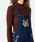 Person wearing a brown ribbed sweater and blue denim overalls with floral embroidery on a white background