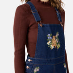 Person wearing a brown ribbed sweater and blue denim overalls with floral embroidery on a white background