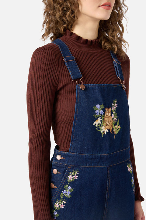 Person wearing a brown ribbed sweater and blue denim overalls with floral embroidery on a white background