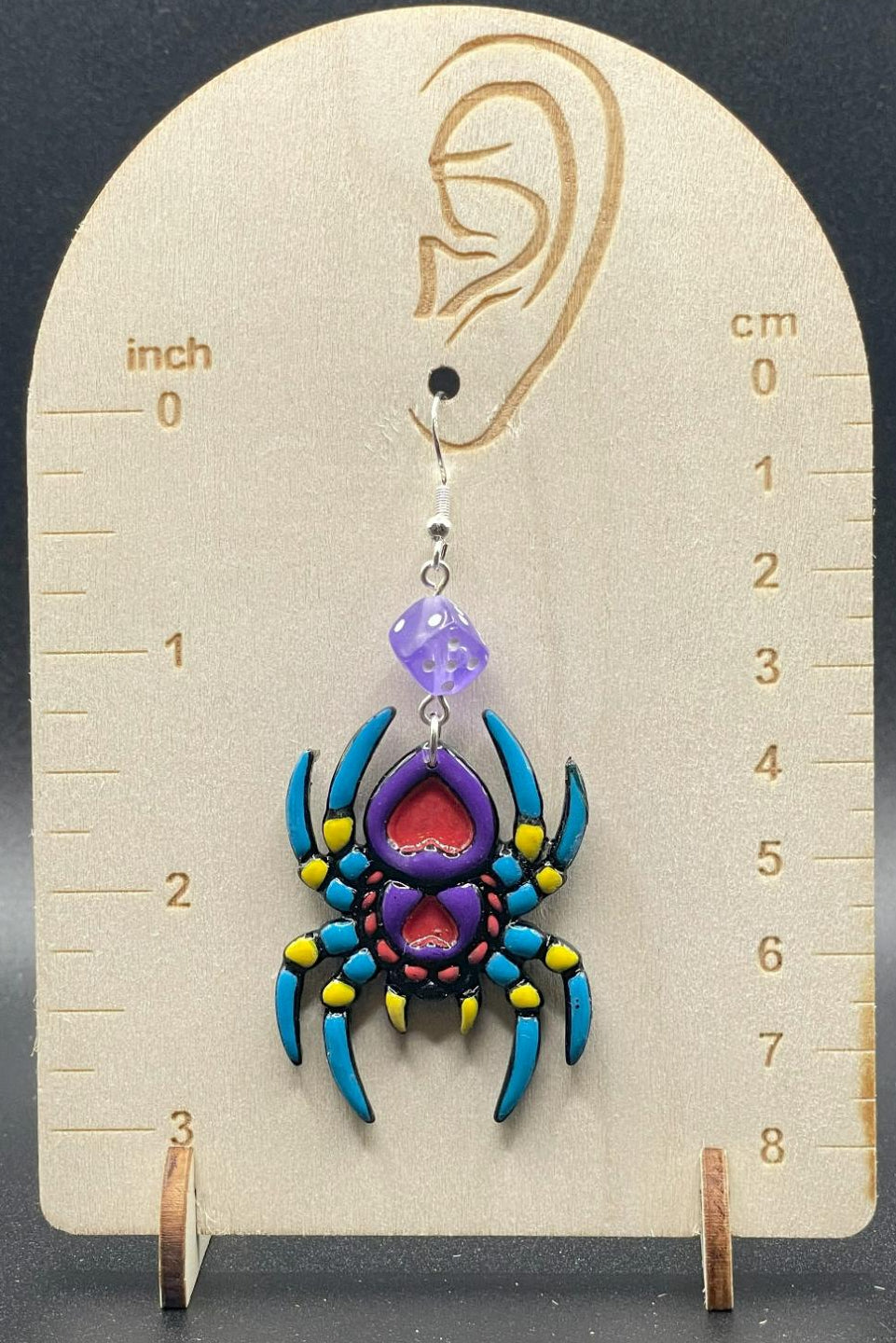 Colorful spider-shaped earring on a wooden display card with measurement scale.