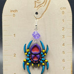 Colorful spider-shaped earring on a wooden display card with measurement scale.