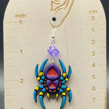 Colorful spider-shaped earring on a wooden display card with measurement scale.