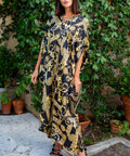 A woman wearing a black and gold baroque-patterned kaftan dress stands outdoors on a stone patio with a lush green garden background.