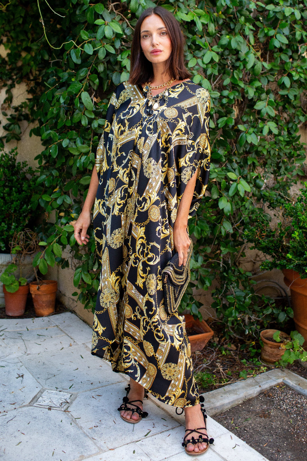 A woman wearing a black and gold baroque-patterned kaftan dress stands outdoors on a stone patio with a lush green garden background.