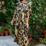 A woman wearing a black and gold baroque-patterned kaftan dress stands outdoors on a stone patio with a lush green garden background.