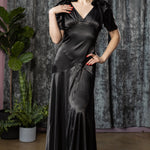Woman wearing a black satin dress standing against a textured curtain backdrop.