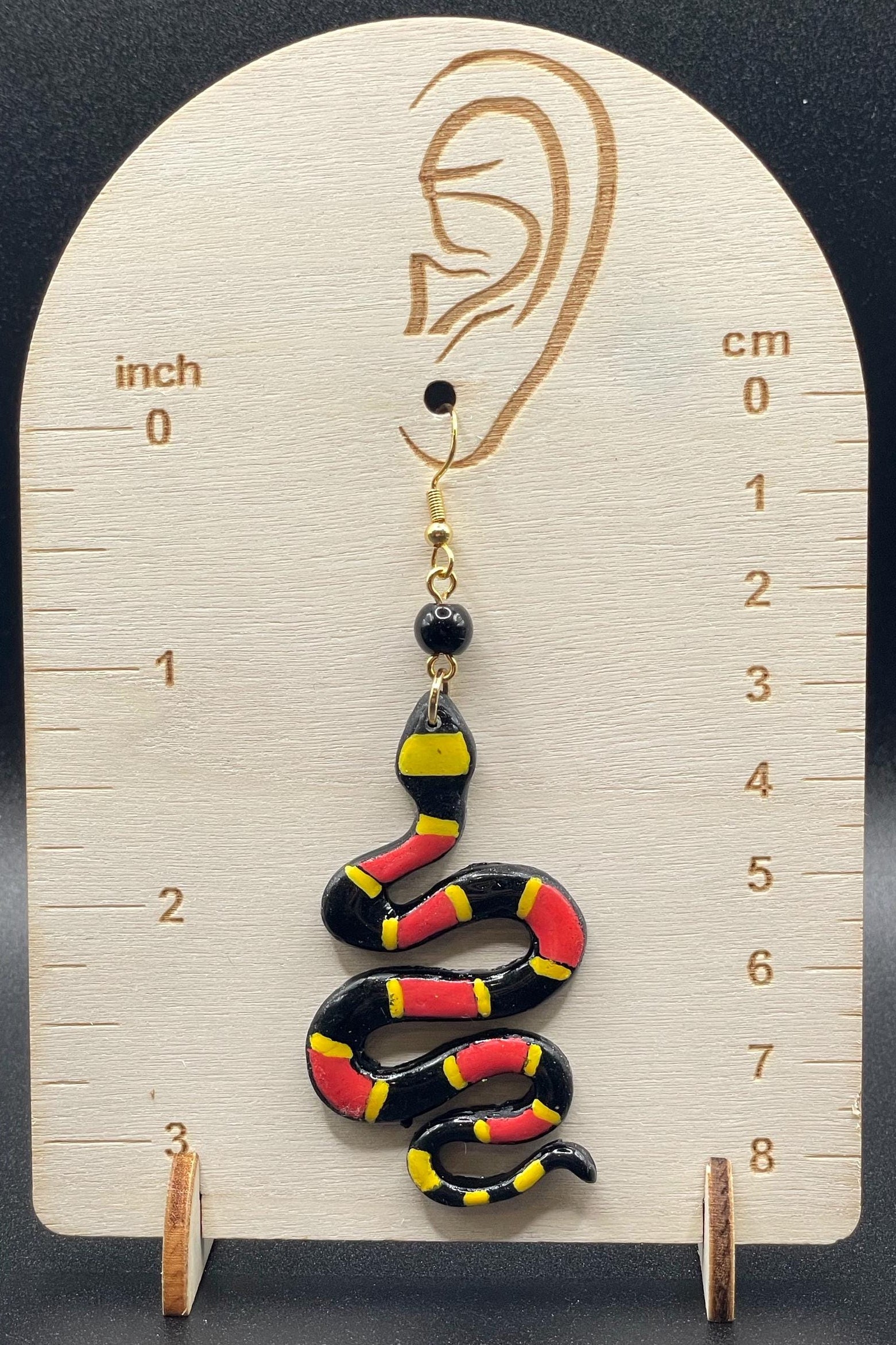 Snake-shaped earring on a wooden display with measurements against a dark background