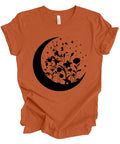 An orange short-sleeve t-shirt with a black graphic of a crescent moon, wildflowers, and stars on the front, displayed on a clean white background.