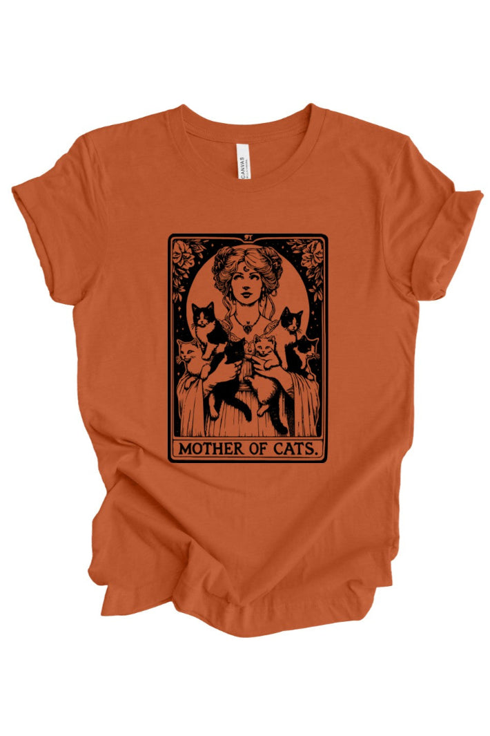 An orange short-sleeve t-shirt with a black graphic print of a woman holding three cats, styled like an Art Nouveau card titled 'MOTHER OF CATS'.